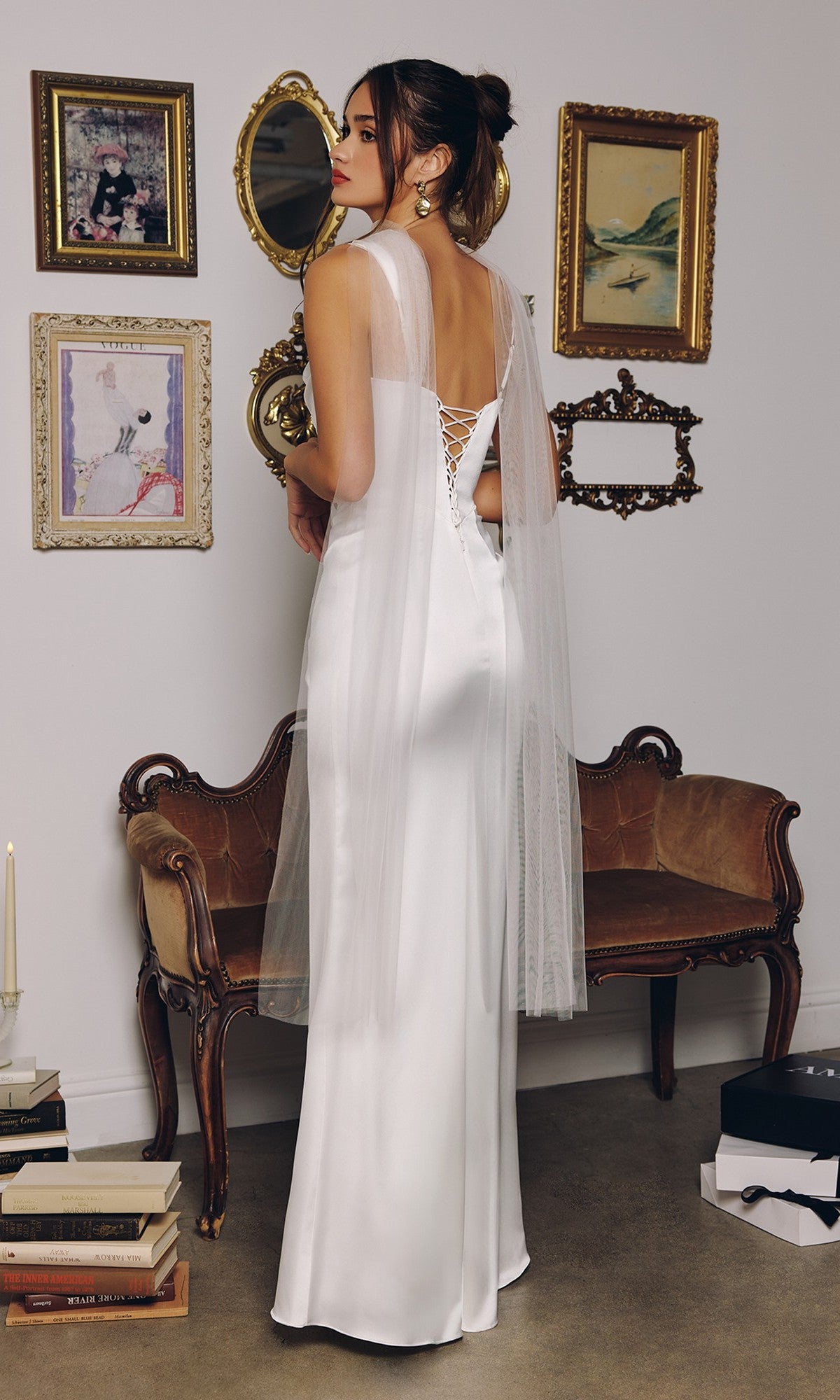 A woman wearing the Amoris T729W Long Wedding Dress, featuring satin white fabric and a sheer cape, stands before a vintage sofa with framed art on the white wall behind her.